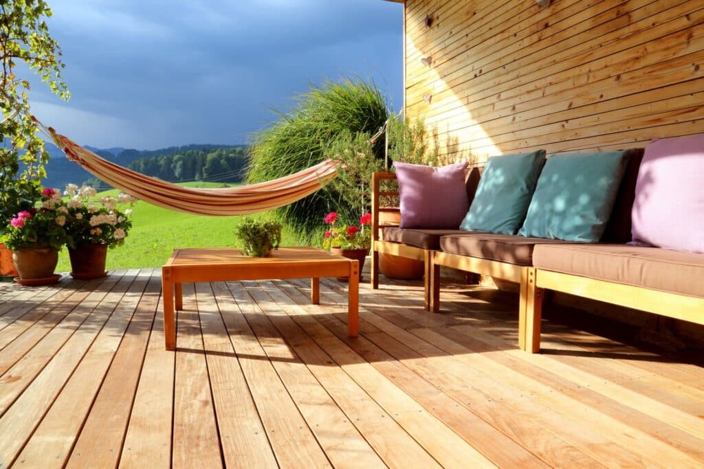 Hängematte und Sitzbereich auf einer Holzterrasse, mit farbigen Kissen und Blick ins Grüne, ideal als gemütliche Ergänzung zu Gartenpavillons für entspannte Stunden im Freien.