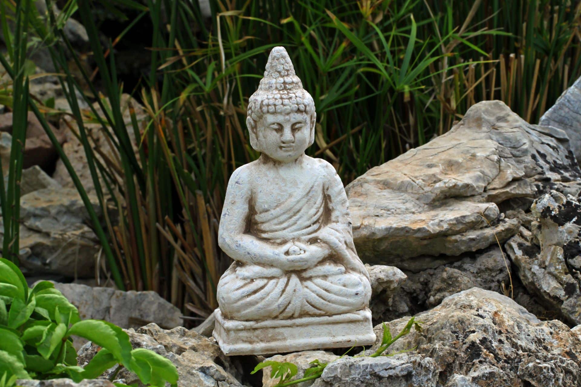 Buddha-Statue, die zwischen Felsen und Gräsern im Garten sitzt, als meditativer Akzent und gestalterisches Element für Ruhebereiche und Gartenpavillons.